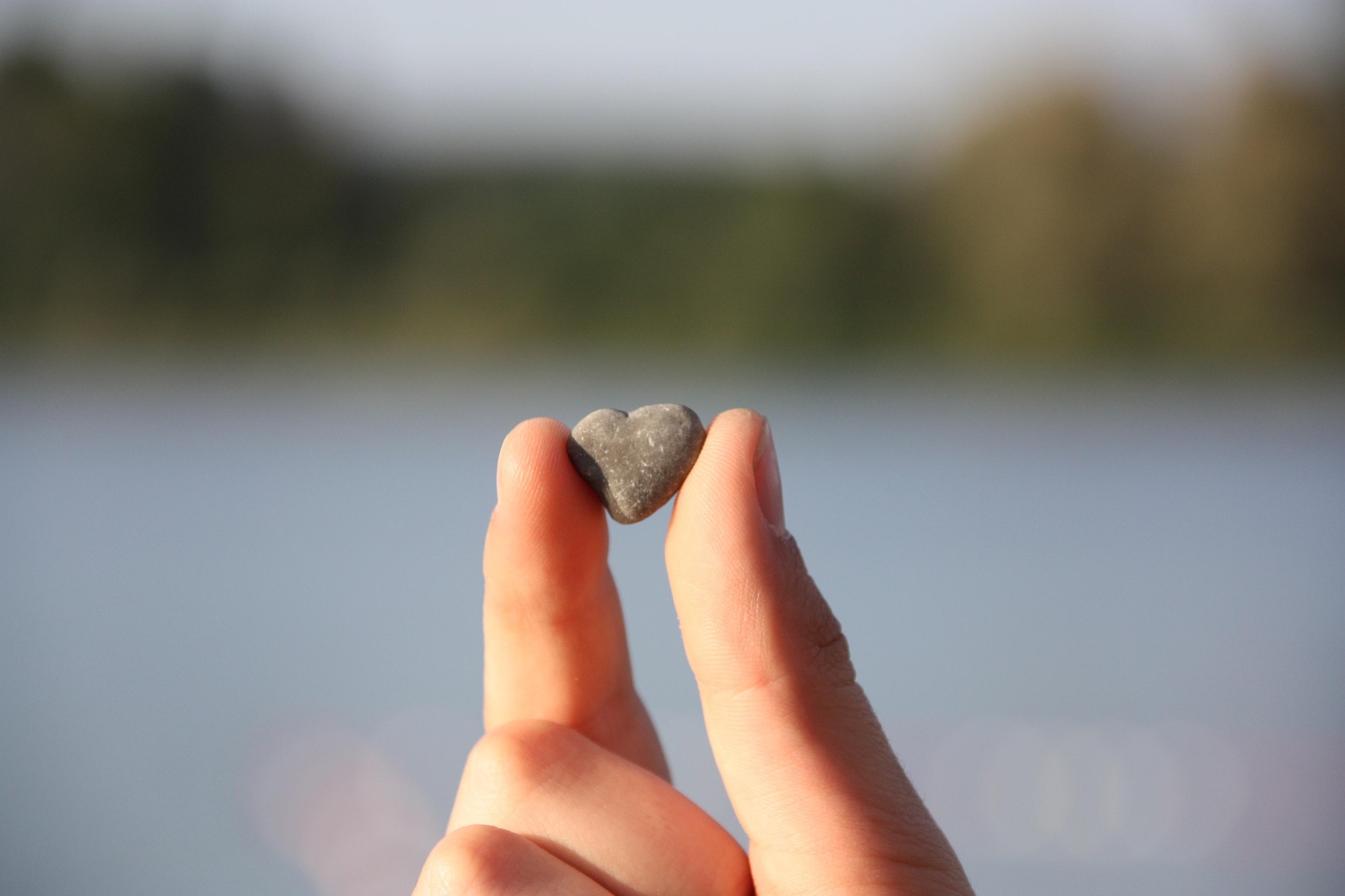 A hand holding a small heart-shaped stone by the water